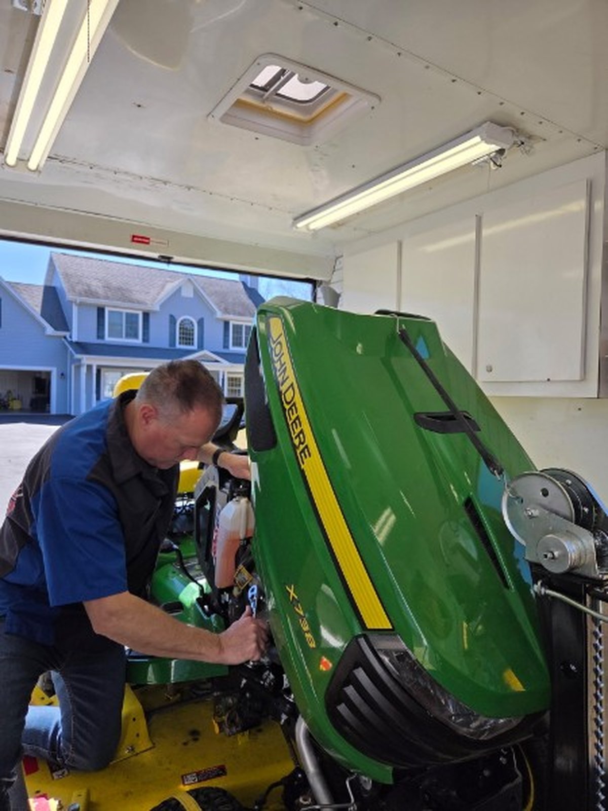 Bill performing hands-on engine repair on a John Deere X738 with the hood raised, neighborhood visible through the open trailer door