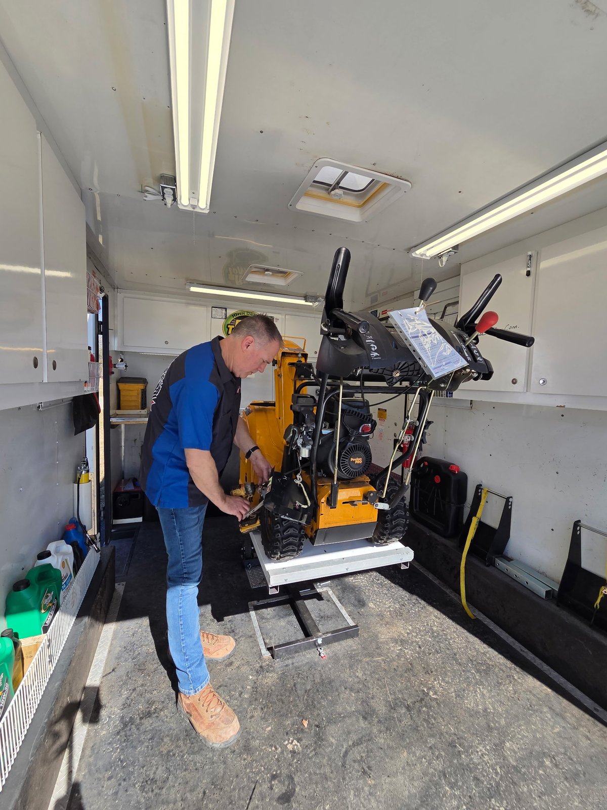 Bill repairing a tracked snowblower on the hydraulic lift, wider angle showing the full mobile trailer interior and tools